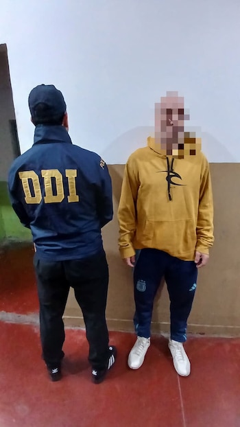 Un agente con una chaqueta que dice DDI permanece de espaldas junto a un hombre con el rostro pixelado frente a una pared