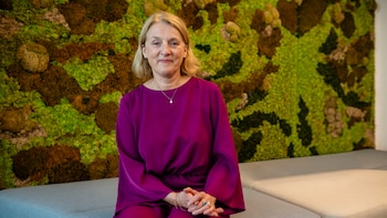 Evelyn Regner, mujer rubia de mediana edad, sentada, sonriendo, con blusa fucsia y collar de perlas, frente a una pared de musgo