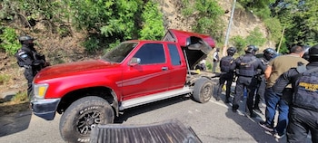 Una camioneta pickup roja modificada en la parte trasera, rodeada por varios agentes de policía y un civil, en una carretera al lado de una ladera con vegetación