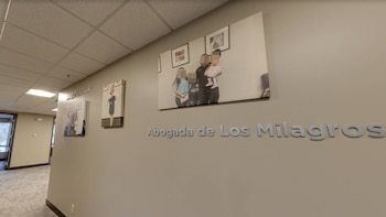 Pasillo de oficina con paredes grises, suelo alfombrado y techo de baldosas. Hay tres cuadros y un letrero con las palabras "Abogada de Los Milagros"