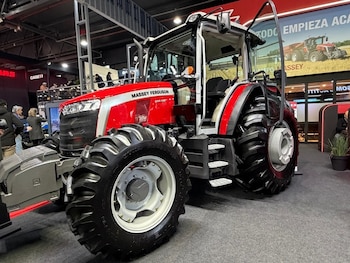 Tractor Massey Ferguson 6M 135 rojo y gris, neumáticos grandes y cabina. Fondo con personas, stands y cartel publicitario de maquinaria agrícola