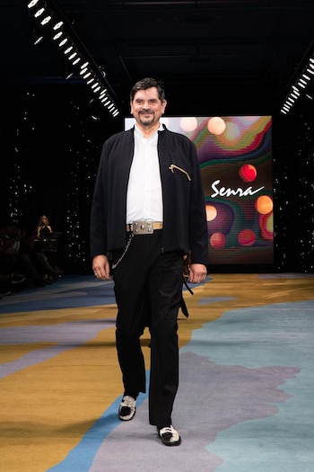 Hombre sonriente en pasarela. Viste chaqueta negra, camisa blanca, pantalones negros, cinturón con hebilla grande y zapatos bicolores. Fondo con pantalla 'Senra'