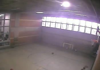 Vista aérea de una cancha interior con canasta de baloncesto y pequeños balones. Grandes ventanas iluminan la pared, y una figura trepa cerca de la canasta