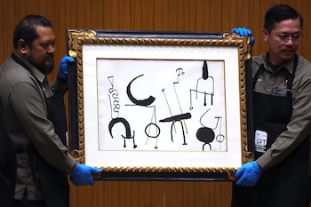 Dos hombres con guantes azules y delantales oscuros sostienen un cuadro con una pintura abstracta de formas negras sobre fondo blanco, enmarcado en dorado y negro