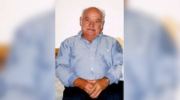 Hombre de edad avanzada con bigote y camisa azul sentado con las manos cruzadas sobre el regazo.