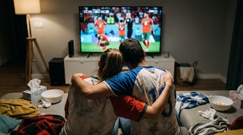 Duas pessoas sentadas costas com costas em um sofá estão se abraçando e assistindo a um jogo de futebol na TV. Lençóis, cobertores e tigelas são fornecidos no quarto.