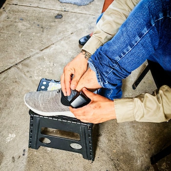 Manos de una persona quitan una etiqueta de seguridad negra de una zapatilla gris, apoyada en un taburete negro, sobre un suelo de concreto