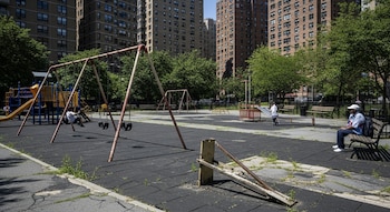 Parque infantil urbano con estructuras de juego, un niño en un columpio y otro en un sube y baja. Un hombre se sienta en un banco, con edificios al fondo.