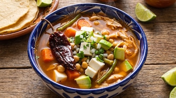 Tazón de cerámica con sopa de pollo, garbanzos, aguacate, queso y un chile seco, acompañado de tortillas de maíz y trozos de limón.
