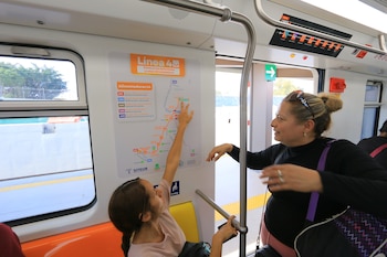 Dos mujeres en el interior de un tren; una señala un mapa de rutas en la pared mientras la otra observa. Se ven asientos anaranjados y ventanas del vagón