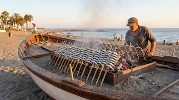 Hombre con gorra cocinando sardinas ensartadas en cañas sobre brasas dentro de una barca en una playa arenosa con el mar de fondo.