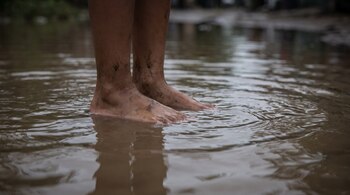 Pies y tobillos descalzos con manchas de barro, parcialmente sumergidos en agua turbia con ondas sobre la superficie.