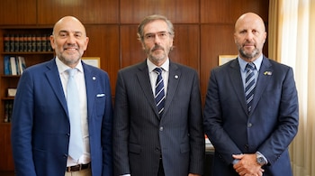 Tres hombres con barba, vestidos con trajes y corbatas, posan de pie en una oficina con paredes de madera y estanterías con libros al fondo.