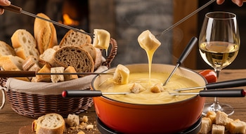 Olla de fondue con queso derretido, tenedores con trozos de pan sumergidos, canasta con pan, copa de vino blanco, mesa de madera, fuego de chimenea en el fondo.