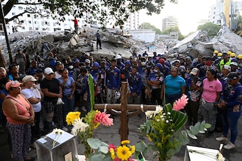 Grupo de personas de pie frente a una cruz de madera adornada con flores de varios colores y montones de escombros de edificios destruidos