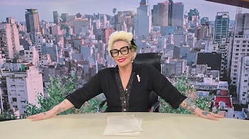 Una mujer con anteojos negros y cabello rubio apoya los brazos en una mesa clara que contiene hojas de papel frente a un fondo urbano.