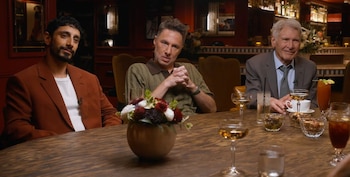 Tres hombres sentados en una mesa de madera oscura en un bar. Hay bebidas como gin, licor y amaro sour, un centro de flores y aperitivos
