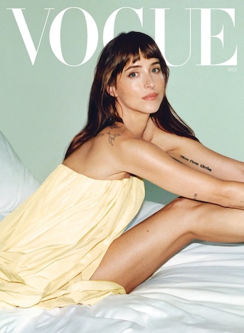 Dakota Johnson sentada en una cama con un vestido amarillo, ante un fondo verde claro y la palabra Vogue en la parte superior