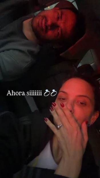 Una mujer sostiene su mano con un anillo cerca de su rostro mientras un hombre sonríe al fondo dentro de un vehículo
