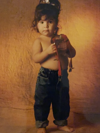 Una niña pequeña con cabello oscuro y rizado, sin camisa, usando pantalones de mezclilla holgados y descalza, sosteniendo tirantes rojos