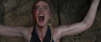Mujer con cabello rubio trenzado, rostro y cuello cubiertos de sangre, boca abierta gritando, brazos extendidos contra una pared rocosa, top negro