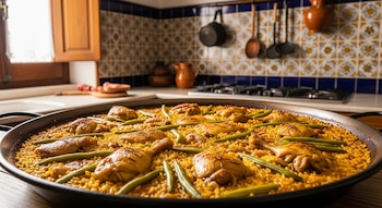 Paella en una paellera grande con arroz dorado, trozos de pollo y conejo, judías verdes y hebras de azafrán. Al fondo, una cocina española tradicional.