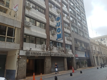 Vista frontal de un rascacielos gris con carteles de 'UOM' y 'Unión Obrera Metalúrgica', muchas unidades de aire acondicionado, y una mujer caminando en la acera de una calle adoquinada