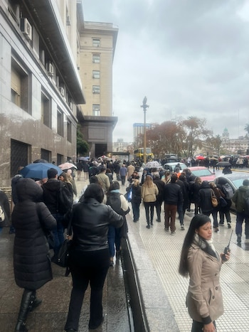 Multitud de personas en la acera frente a un edificio gubernamental en un día nublado y lluvioso. Algunas personas usan paraguas. Una mujer sostiene una radio