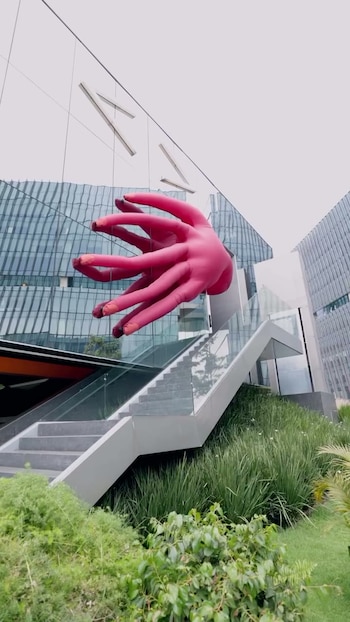 Edificio moderno de cristal con letras "AY" y una escultura de mano inflable rosa. Se observan escaleras de hormigón con barandales de cristal y plantas