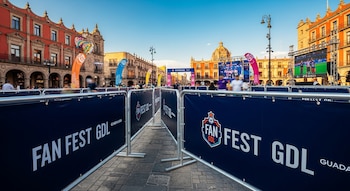 Vista frontal de vallas azules con logos "FAN FEST GDL" en una plaza con edificios históricos coloniales de fondo, pantallas gigantes y personas difuminadas.