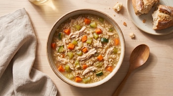 Un plato hondo con sopa de avena, pollo, zanahoria, calabacín y apio sobre una mesa de madera clara, junto a una cuchara de madera, una servilleta y pan.