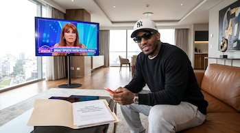 Jefferson Farfán, con gorra blanca y lentes de sol, sentado en un sofá, sostiene un celular. Una carpeta abierta con documentos está en una mesa. Una TV muestra a Magaly Medina.