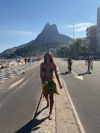 Calu Rivero camina descalza por una acera de adoquines en un día soleado, vistiendo un bikini y pareo verde y amarillo. Al fondo, playa, mar y montañas