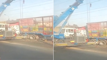 Una grúa azul con su brazo extendido y gancho visible está junto a un camión blanco con cabina roja que transporta jaulas de colores en una carretera