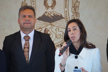 Un hombre y una mujer. La mujer habla en un micrófono y sostiene un teléfono. El hombre, de traje y corbata estampada, mira al frente. Fondo con emblema