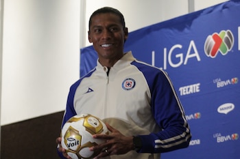 Joel Huiqui sonríe sosteniendo un balón de fútbol Voít, vestido con una chaqueta blanca y azul del Cruz Azul, frente a un fondo azul de la Liga MX