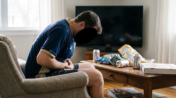 Um homem com uma camisa de futebol está sentado em uma cadeira, segurando a barriga, cercado por salgadinhos e recipientes de comida vazios em uma mesa de centro.