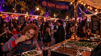 Un hombre disfrazado come tacos frente a recipientes con guisados en un mercado nocturno decorado con guirnaldas y calabazas de Halloween