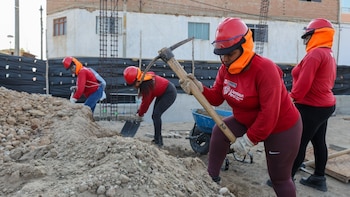 Cuatro trabajadoras con cascos rojos, lentes de protección y camisetas rojas usan picos y palas sobre montículos de tierra
