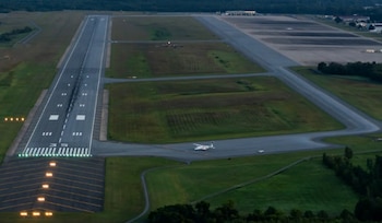Fotografía aérea de una pista de aeropuerto asfaltada con marcas blancas, luces verdes y amarillas, un avión blanco en un taxiway y un auto blanco