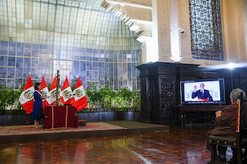 Salón con mujer de pie, cinco banderas de Perú, mesa con paño rojo, cruz. Pantalla de televisión muestra a hombre levantando mano. Persona sentada de espaldas