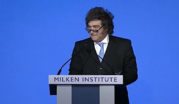 Javier Milei, con traje oscuro, camisa blanca y corbata azul, habla desde un podio blanco con el logo 'MILKEN INSTITUTE' sobre un fondo azul liso