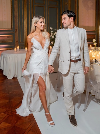 Pareja, ella con vestido de novia blanco y él con traje claro, caminan de la mano en un salón elegante con paredes de madera y mesas con velas