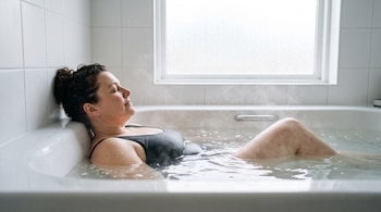Una mujer con traje de baño negro recostada en una bañera llena de agua, junto a una pared de azulejos blancos y una ventana.