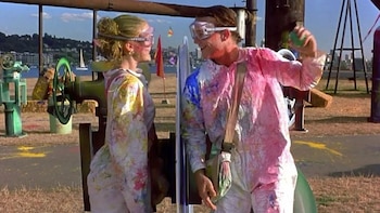 Heath Ledger y Julia Stiles, con trajes protectores manchados de pintura y gafas transparentes, se miran y sonríen en un área abierta frente a un lago