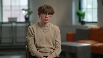 Un hombre joven con cabello castaño desordenado, gafas negras y suéter beige claro, sentado en una silla. El fondo es un interior borroso con un sofá naranja