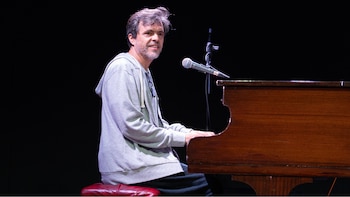 Un hombre con sudadera gris está sentado al piano tocando, con un micrófono en un soporte frente a él y un fondo oscuro