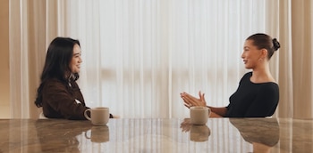Dos mujeres sentadas en una mesa brillante con dos tazas, conversando. La mujer de la derecha, con moño, gesticula; la de la izquierda, con cabello largo, sonríe