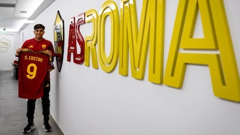 Hombre sonriente, Santiago Castro, sostiene una camiseta de fútbol roja con el número nueve y su nombre. Detrás, letras de AS Roma en una pared blanca