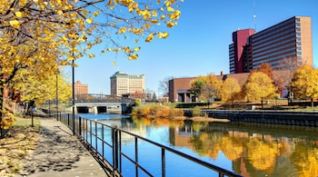 Vista de un río flanqueado por un sendero con barandilla, árboles de hojas amarillas de otoño y edificios urbanos bajo un cielo azul claro.